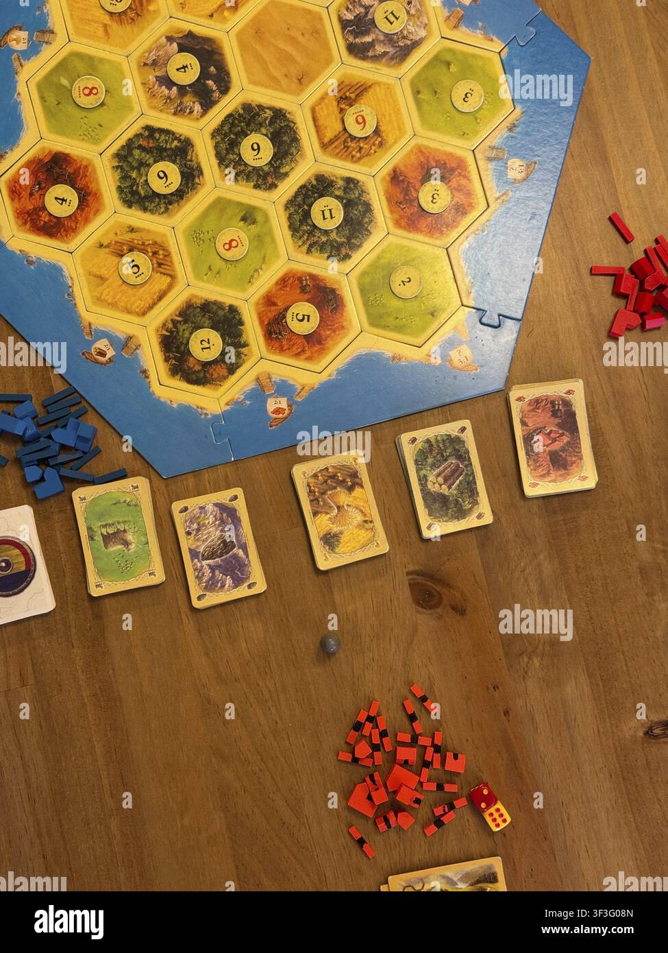 Configurazione del tabellone di Catan su tavolo di legno, carte risorse sparse e insediamenti colorati con gettoni rossi e blu, giocatori che preparano le mosse Foto Stock