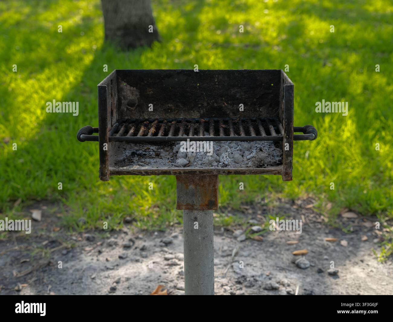 Un barbecue metallico stazionario contenente cenere di carbone bruciata poggia saldamente su un palo di acciaio spesso all'interno di un'area ricreativa all'aperto designata. Foto Stock