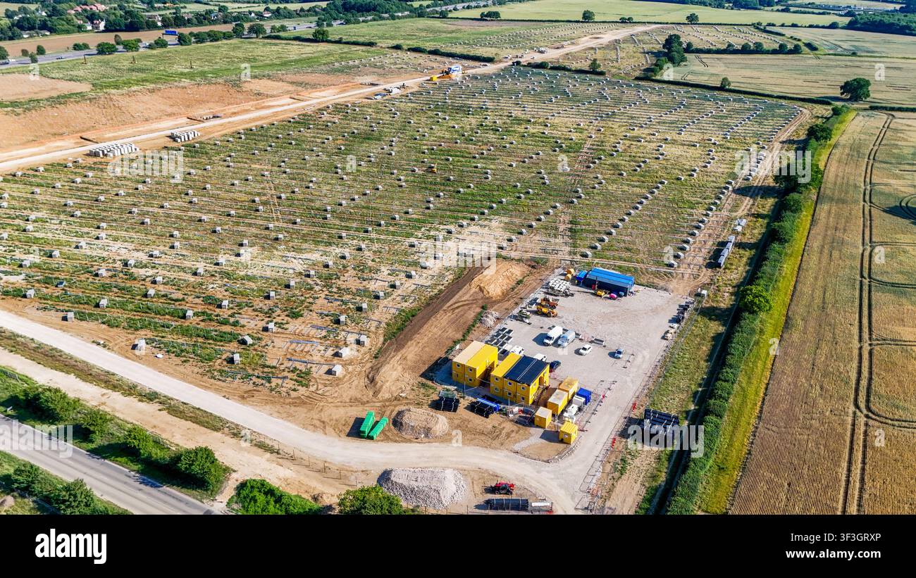 Vista aerea del cantiere di costruzione di una fattoria solare con file di fondamenta in cemento, container edili e macchinari pesanti nel paesaggio rurale di Gre Foto Stock