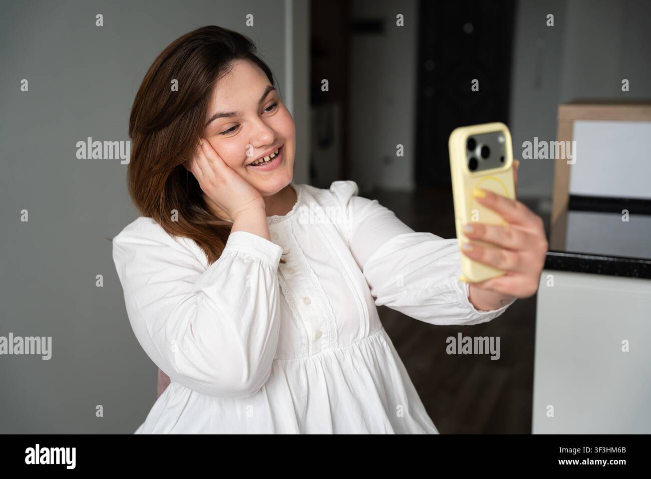 Giovane donna che sorride mentre tiene uno smartphone per una videochiamata in un ambiente domestico moderno. Marketing di influencer sui social media e branding personale. Foto Stock