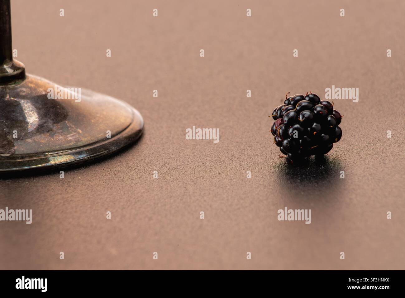 Un colpo minimalista orizzontale che si concentra su una singola more matura appoggiata su una superficie scura accanto all'elegante stelo di un calice di metallo. La composizione Foto Stock