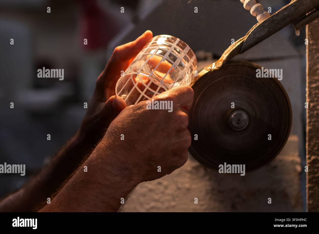 Le mani esperte di un soffiatore di vetro incidono la superficie di un bicchiere di cristallo soffiato su una mola rotante in ghisa bagnata, creando un motivo decorativo preciso Foto Stock