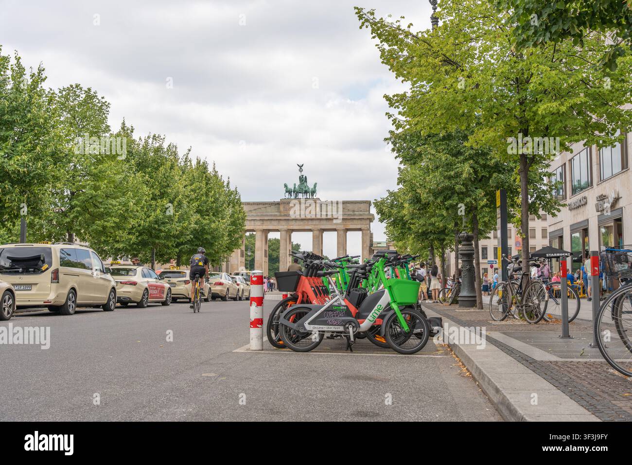 Berlino, Germania, 18 luglio 2026. Biciclette a noleggio e taxi fiancheggiano Unter den Linden di fronte alla Porta di Brandeburgo. I ligustri del genere Tilia incorniciano questo autobus Foto Stock