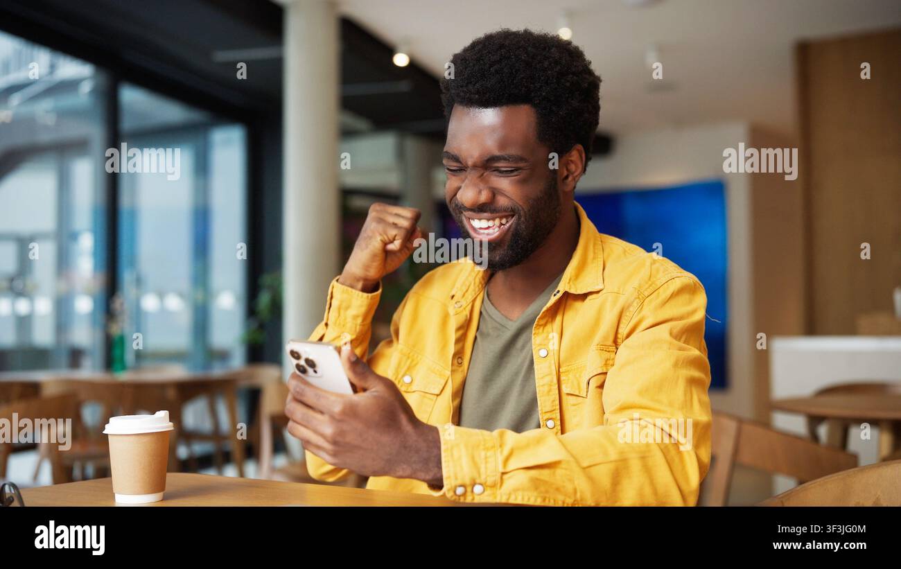 Uomo afroamericano felice che celebra il successo guardando il smartphone in un caffè. Uomo che alza il pugno e ride con entusiasmo mentre controlla buone notizie Foto Stock