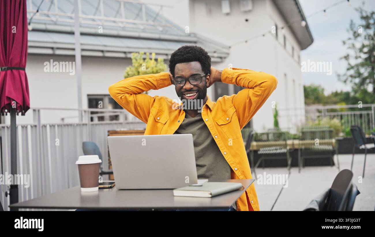 Affascinante uomo afroamericano che alza le mani sorpreso mentre lavora su un laptop all'aperto. Uomo seduto a un tavolo con una tazza di caffè e un taccuino, che reagisce Foto Stock