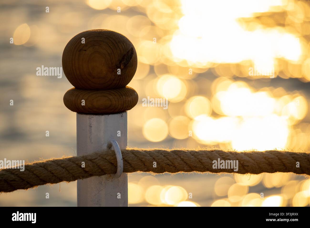 Primo piano di un palo in legno del molo con una ringhiera in corda contro un bokeh d'acqua dorata al tramonto Foto Stock