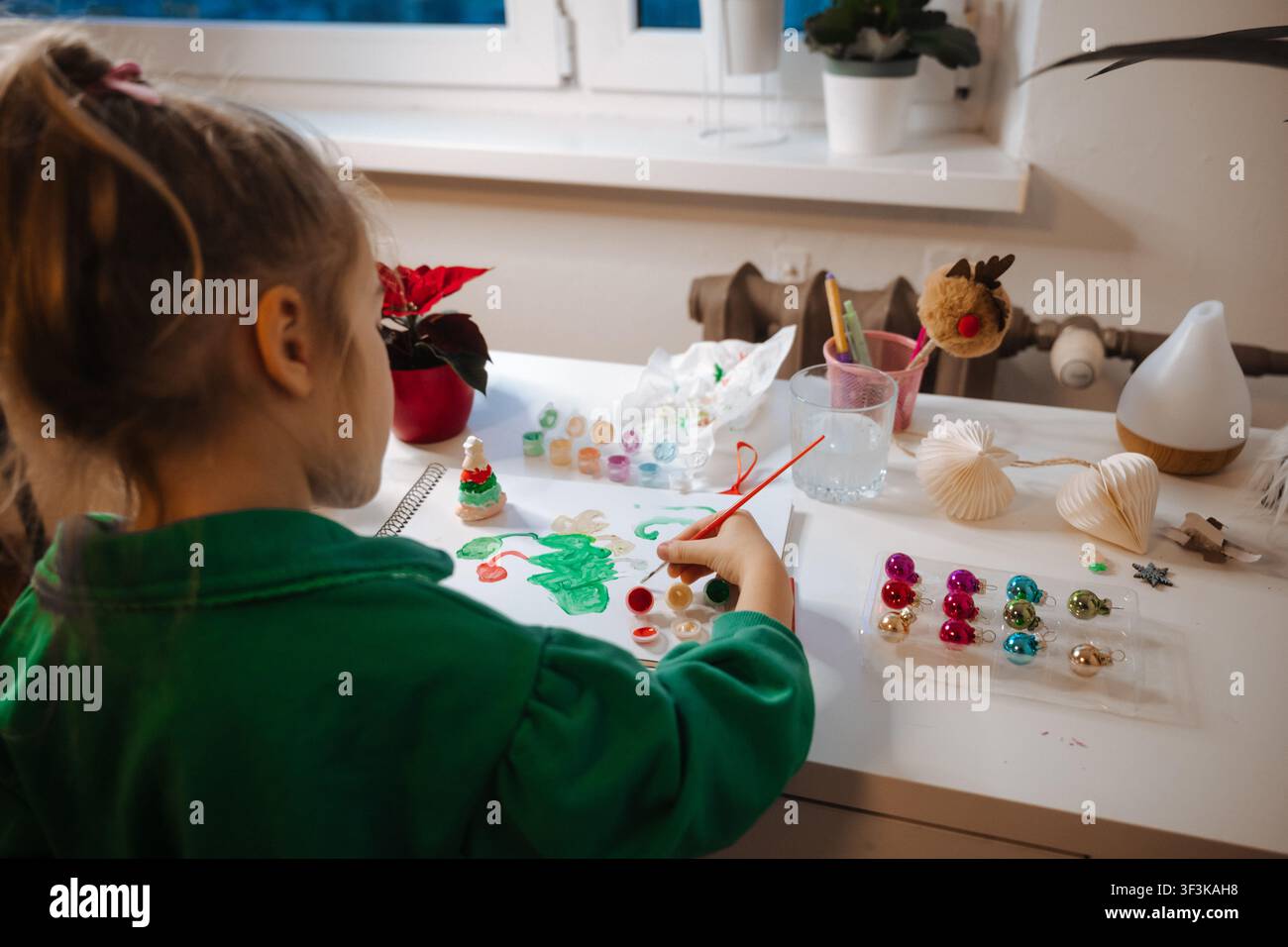 Un bambino dipinge decorazioni seduto a un tavolo in uno spazio interno accogliente durante il giorno Foto Stock