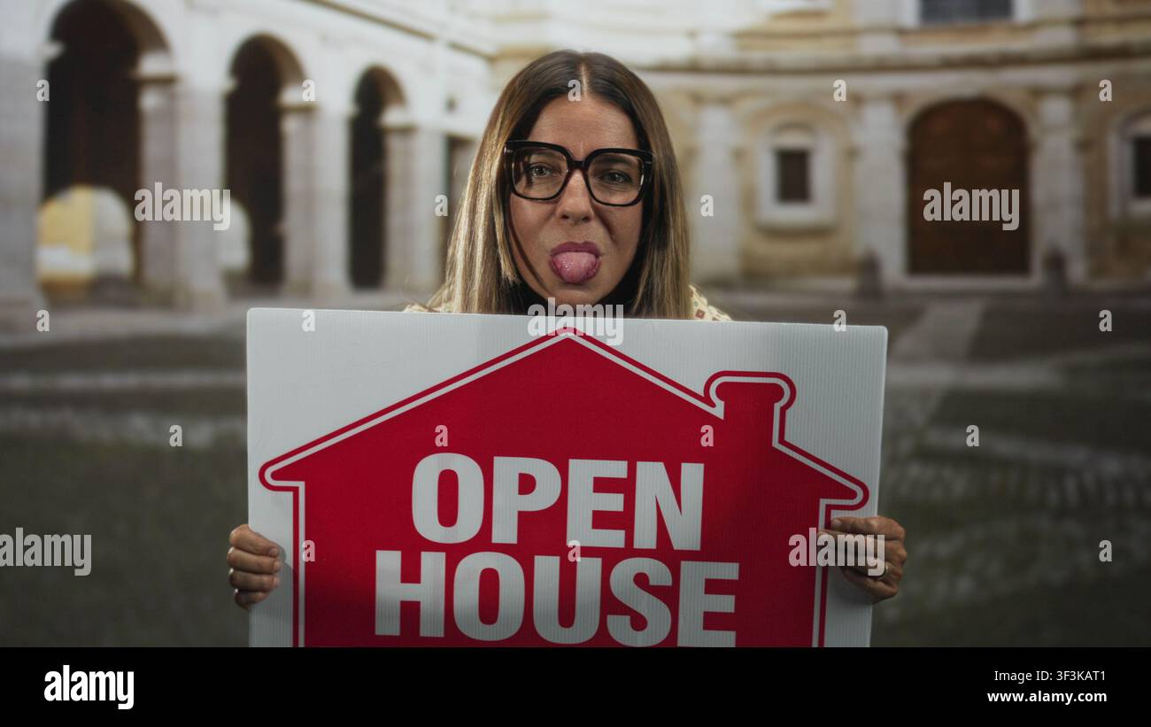 Donna che indossa occhiali con un cartello rosso aperto e che sporge la lingua, un cartello con le mani davanti agli archi del cortile dell'edificio; promo giocoso Foto Stock