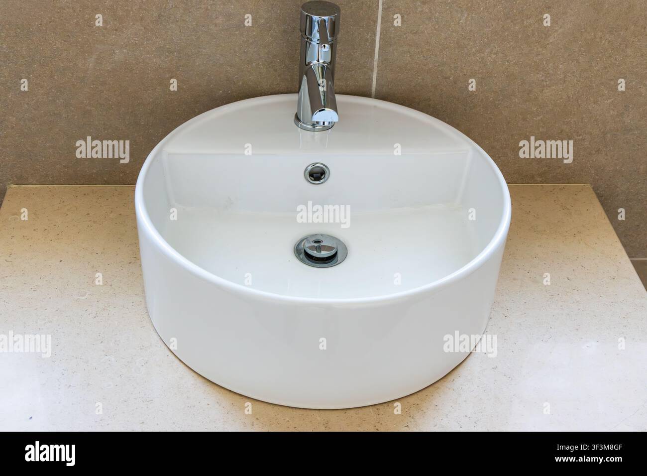 Un lavandino da bagno contemporaneo e pulito con un bacino bianco rotondo, un rubinetto cromo elegante e un piano di lavoro beige strutturato Foto Stock