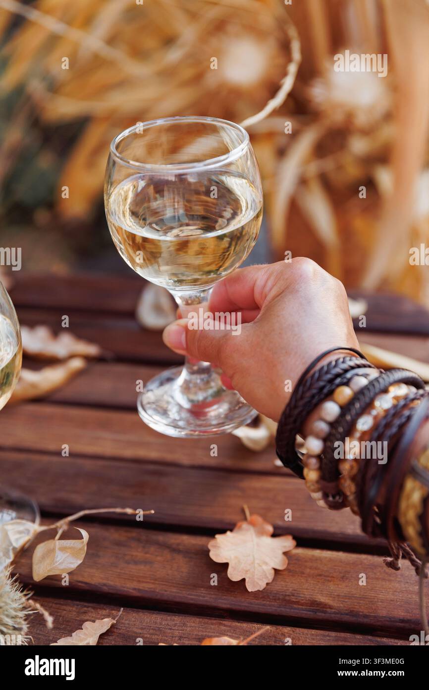 Mano femminile con braccialetti bohémien che tiene un bicchiere di vino bianco sopra un tavolo di legno rustico circondato da foglie autunnali e colori caldi stagionali Foto Stock
