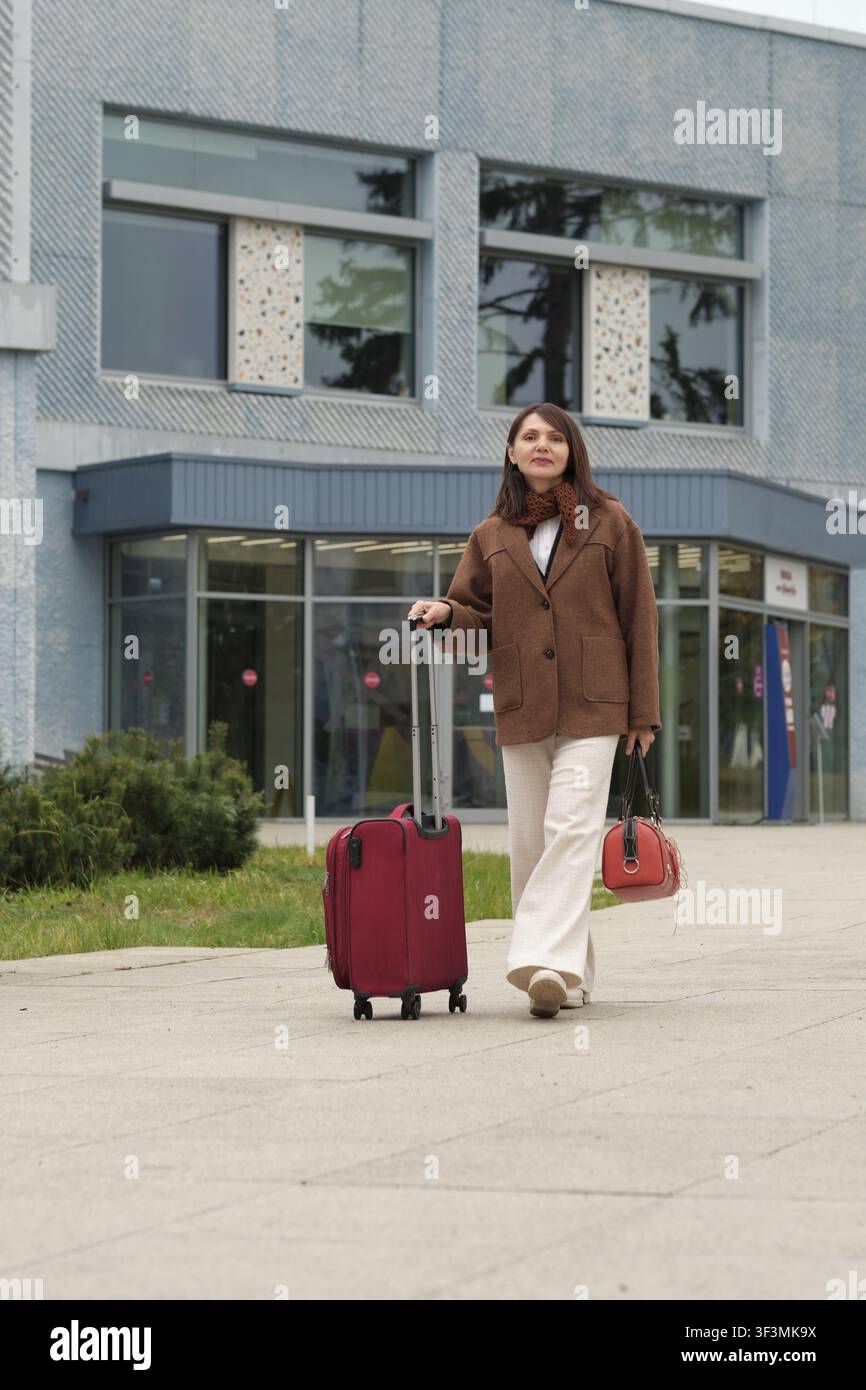 Un viaggiatore con un caldo abbigliamento autunnale si erge su un marciapiede con la sua valigia. La composizione evoca libertà, movimento e un'estetica moderna. Foto Stock
