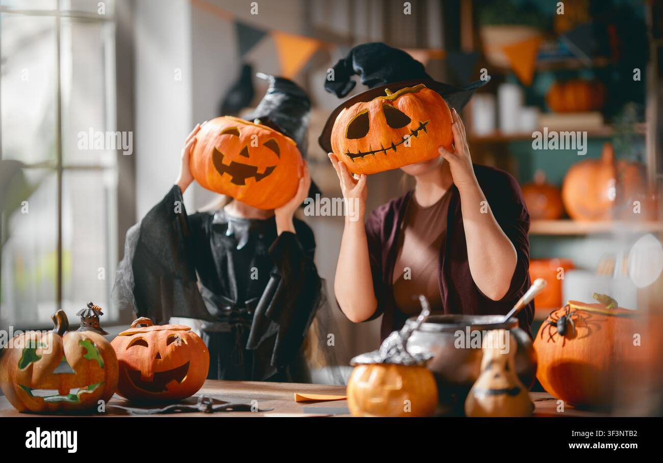 Gioiosa celebrazione familiare di Halloween con madre e figlia che indossano cappelli da strega e tengono zucche intagliate sopra i loro volti. L'interno accogliente della casa i Foto Stock