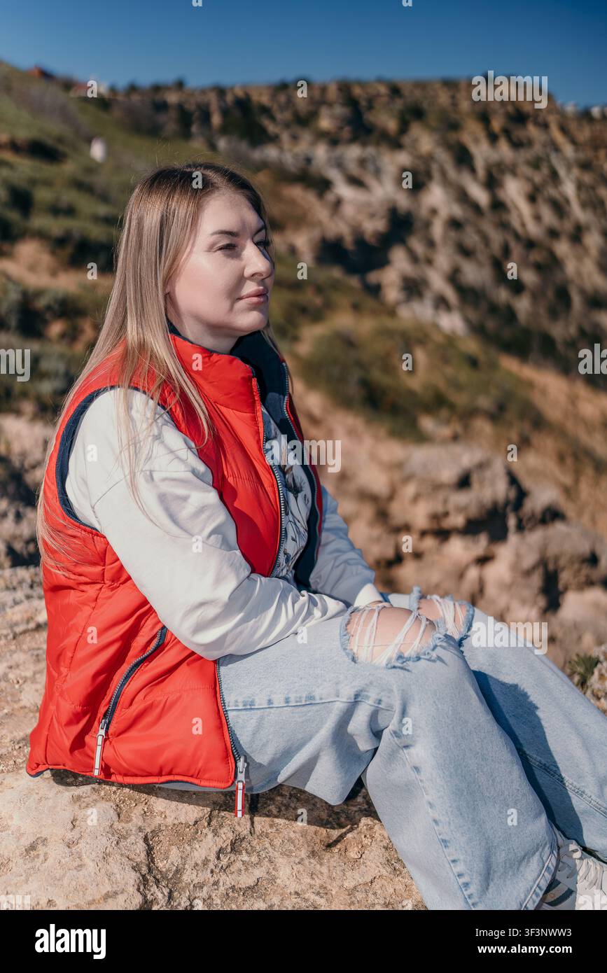 Donna che fa escursioni in montagna si siede su una cresta rocciosa indossando un giubbotto rosso e jeans strappati, godendosi la natura in una giornata soleggiata e luminosa Foto Stock
