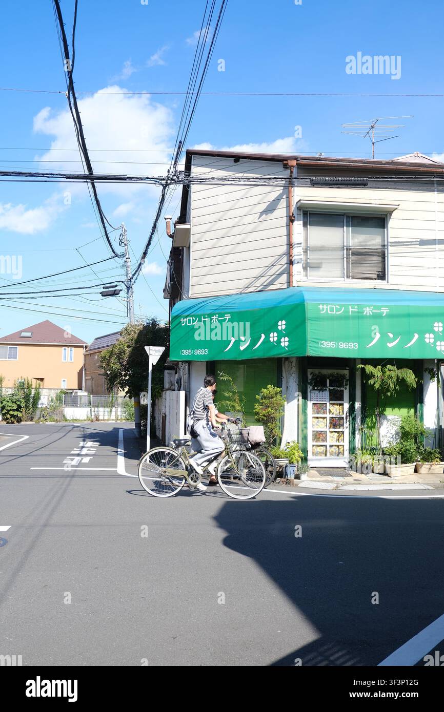 Una persona in bicicletta passa davanti a un salone di bellezza locale con una tenda verde in un angolo di strada soleggiato in un quartiere giapponese sotto un cielo blu terso. Foto Stock