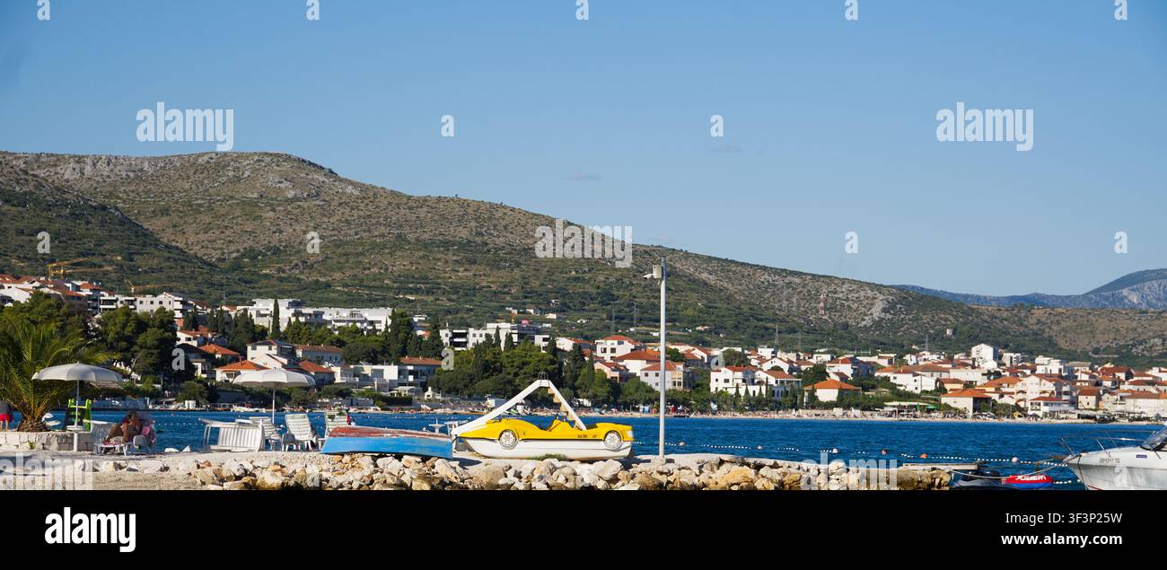 Barca a pedali gialla e sedie da spiaggia sulla riva con una città di resort costiera e colline sullo sfondo. Foto Stock