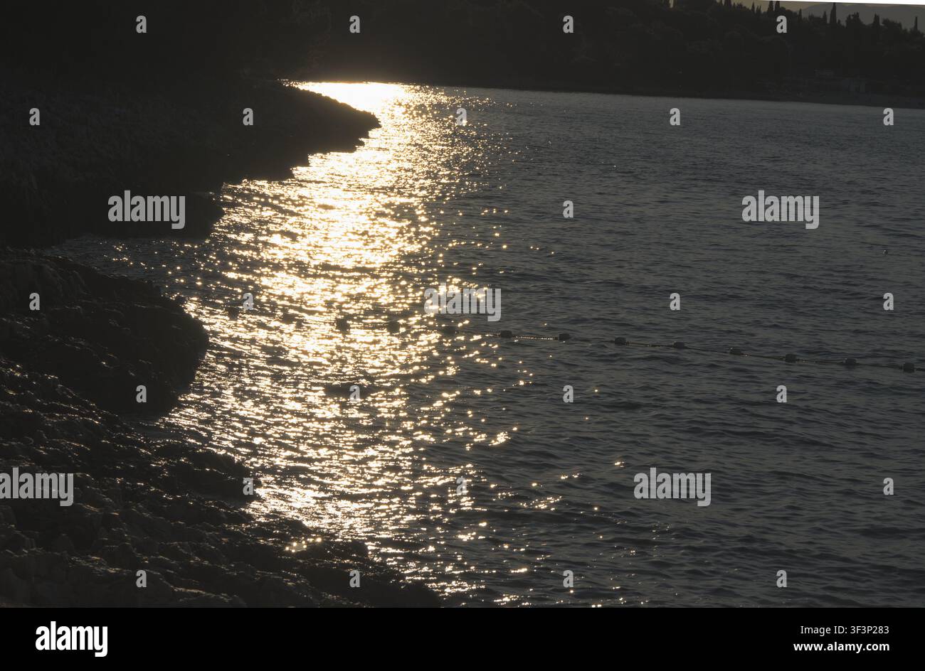 Luce dorata del sole che brilla sulle increspature del mare contro la drammatica silhouette di una riva rocciosa. Foto Stock