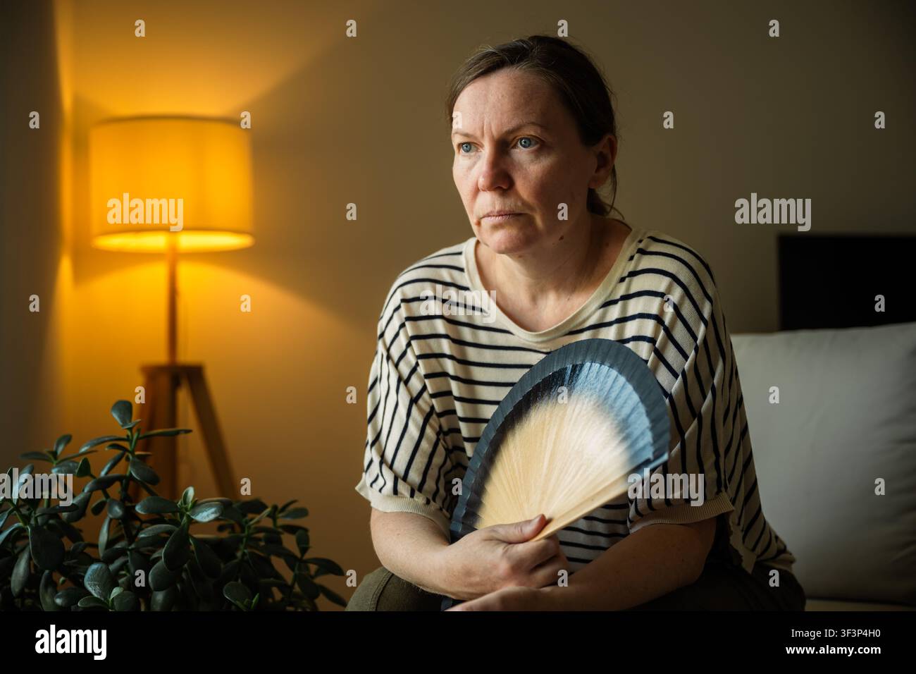 Donna di mezza età che si rinfresca con un ventaglio portatile durante una calda serata estiva a casa. Concetto di ondata di caldo, disagio e mancanza di aria condizionata. Foto Stock