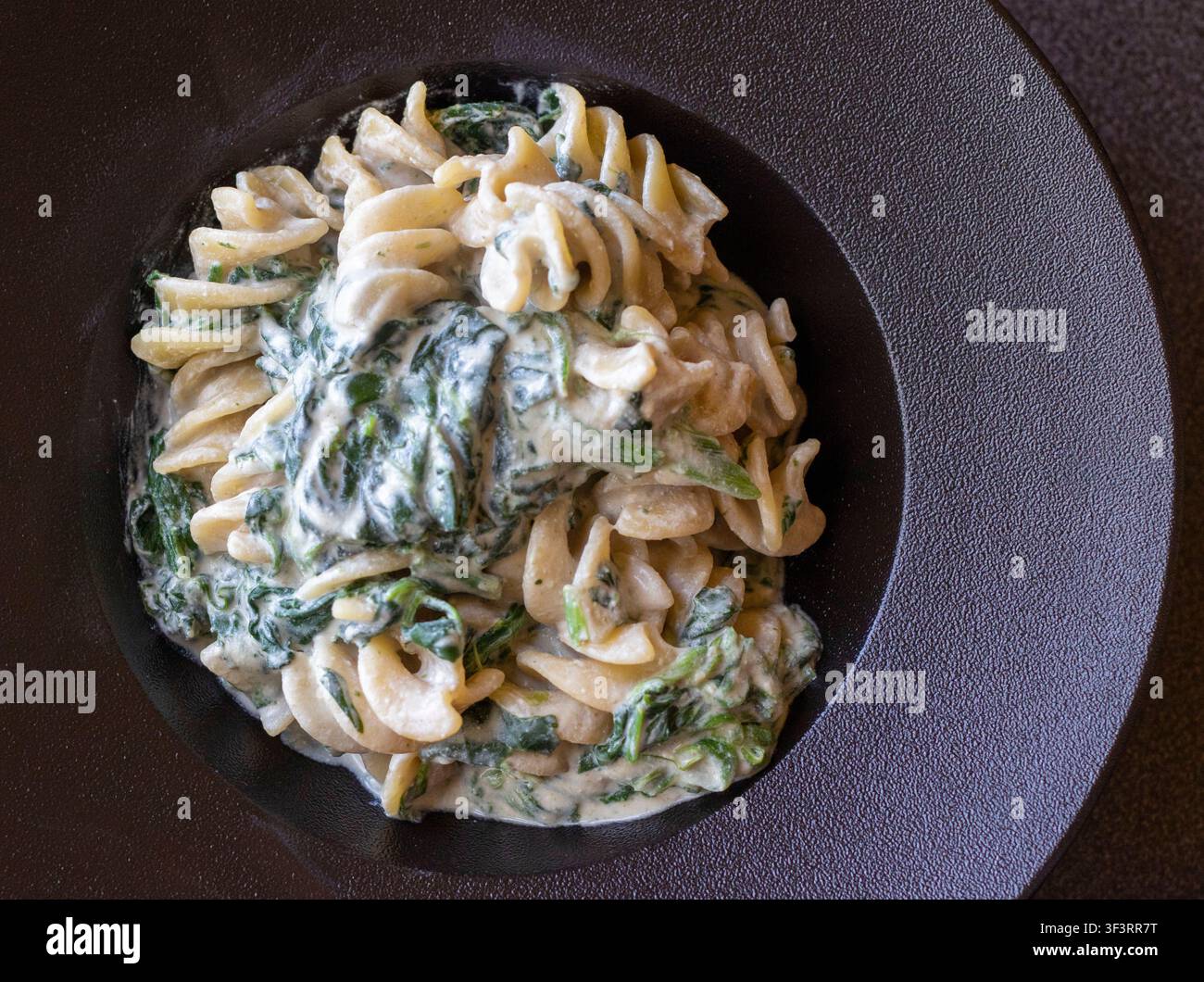 Vista dall'alto di pasta fusillotti con spinaci appassiti in salsa cremosa di acciughe saporite su un piatto di ceramica scura, cucina casalinga italiana autentica Foto Stock