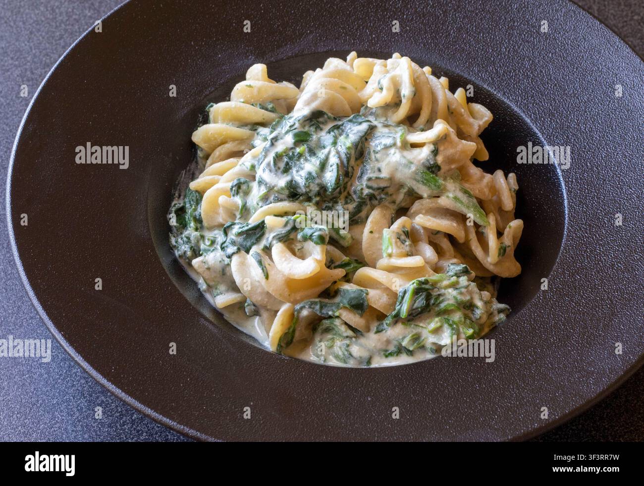 Vista dettagliata di pasta fusillotti con spinaci appassiti in salsa cremosa di acciughe saporita su un piatto di ceramica scuro, cucina casalinga italiana autentica Foto Stock
