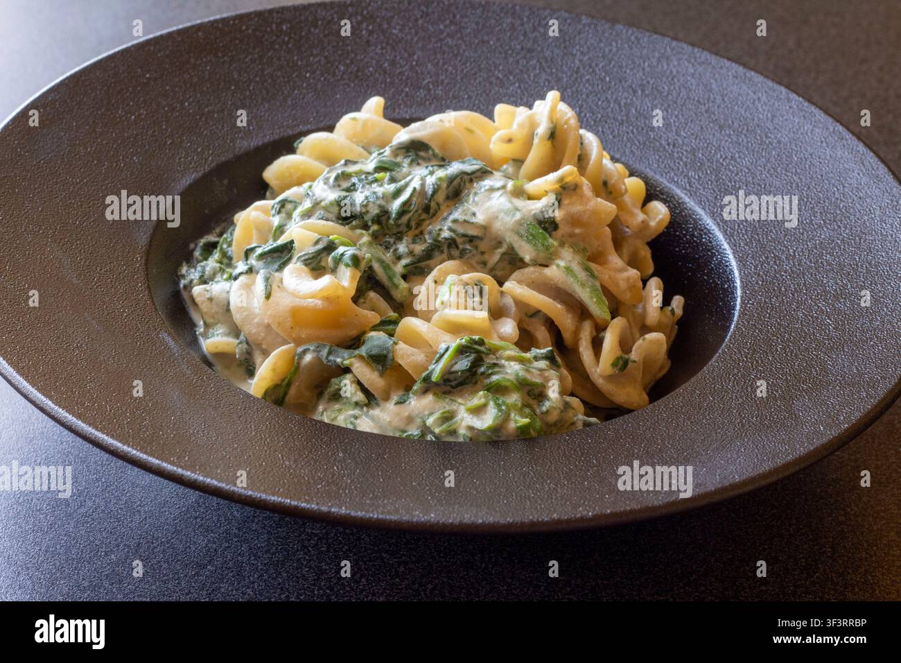 Vista angolata di pasta fusillotti con spinaci appassiti in salsa cremosa di acciughe saporita su un piatto di ceramica scura, cucina casalinga italiana autentica Foto Stock