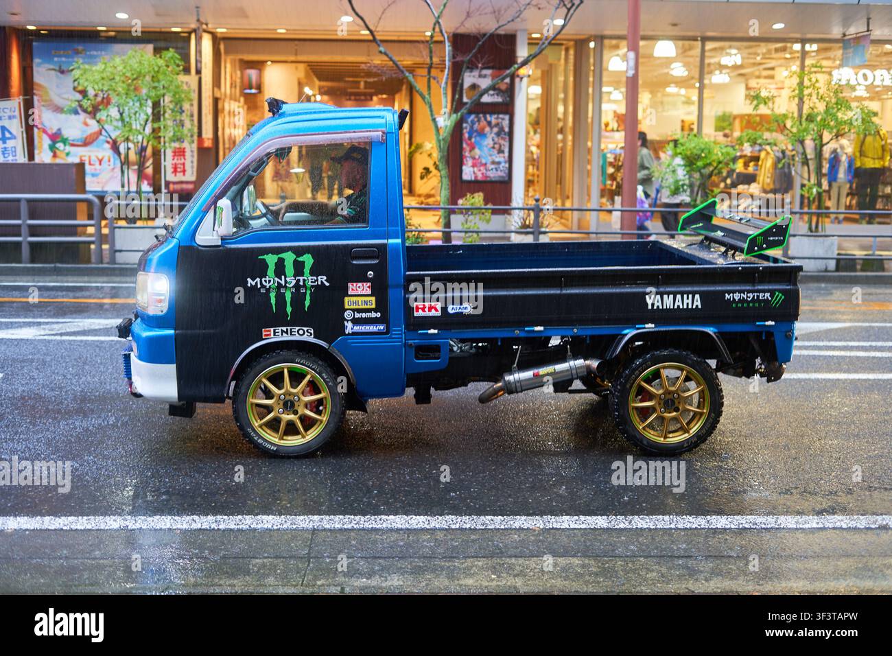 Un mini camioncino Kkei giapponese modificato blu e nero parcheggiato su una strada urbana bagnata davanti ad edifici commerciali. Il veicolo è dotato di una lega dorata Foto Stock