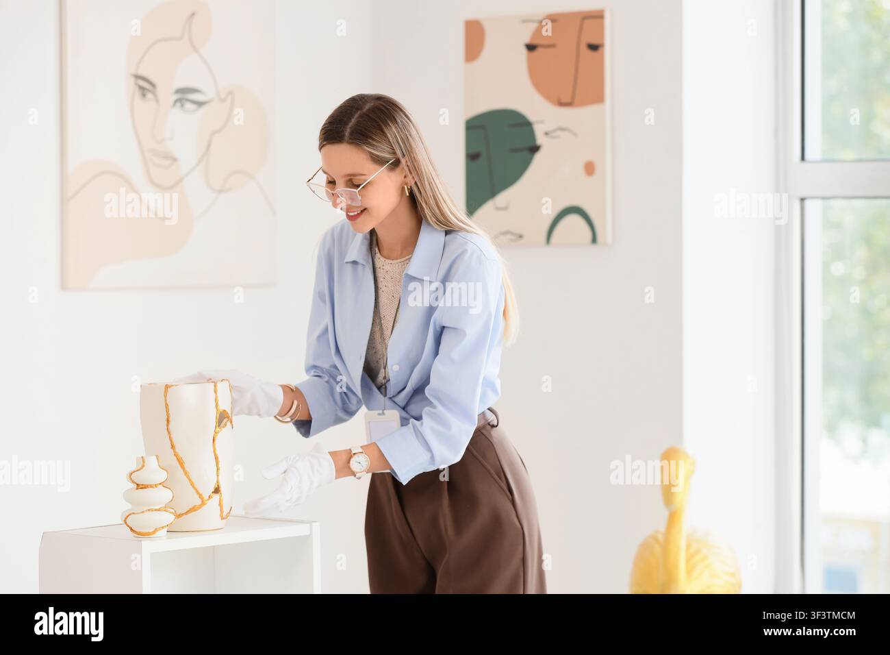 Giovane curatrice di galleria d'arte con guanti bianchi che sistema con cura la ceramica kintsugi su una mensola espositiva Foto Stock
