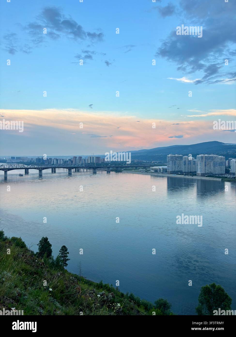 KRASNOYARSK, RUSSIAN - LUGLIO 2026: Vista panoramica del fiume Enisej con ponte stradale, skyline della città e montagne al tramonto sereno, Siberia - Immagine stock catturata con smartphone