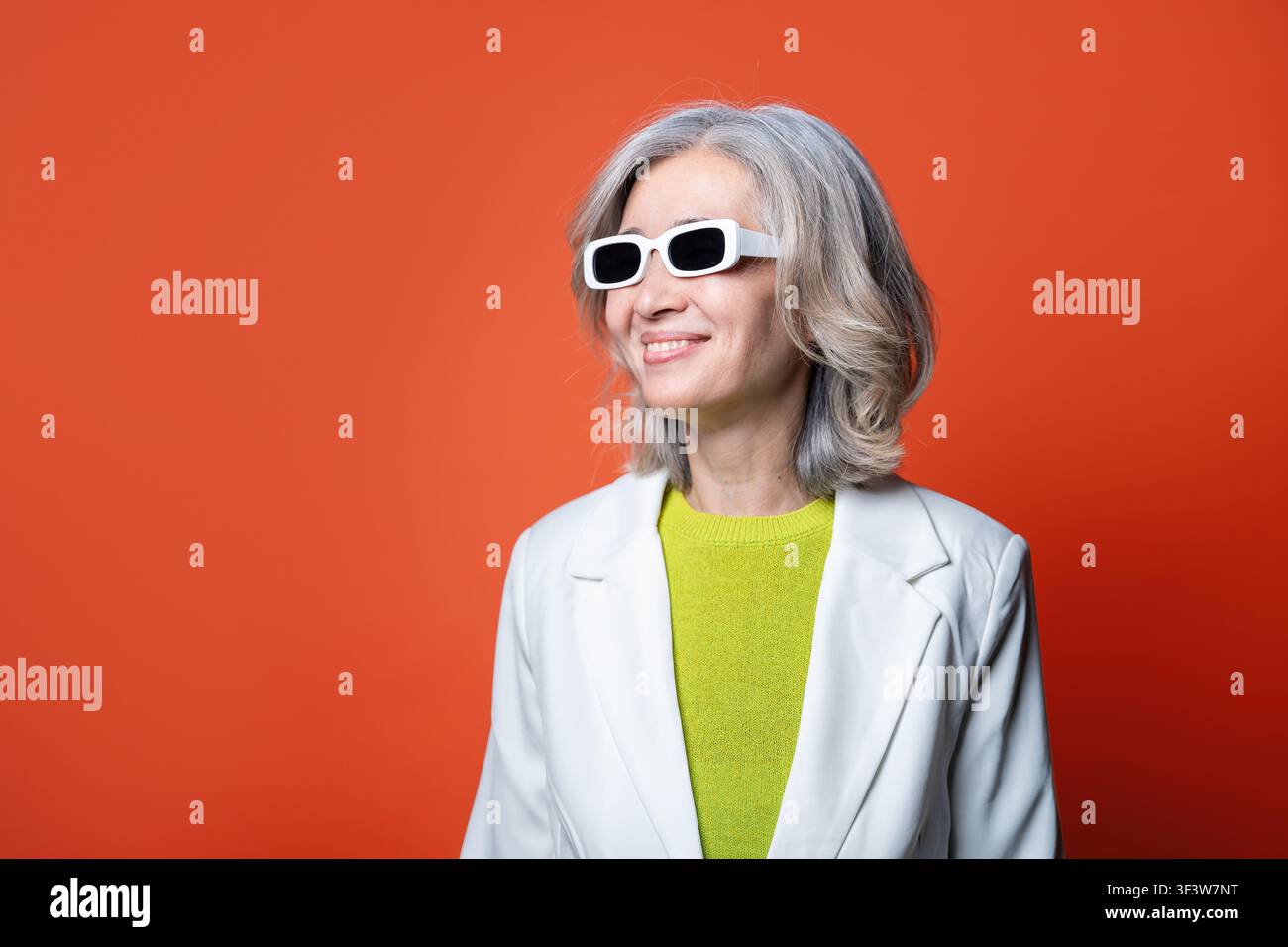 Ritratto di studio di una donna asiatica anziana con capelli grigi che indossa occhiali da sole bianchi, un blazer e un maglione verde lime, sorridendo e guardando di lato contro uno sfondo luminoso Foto Stock