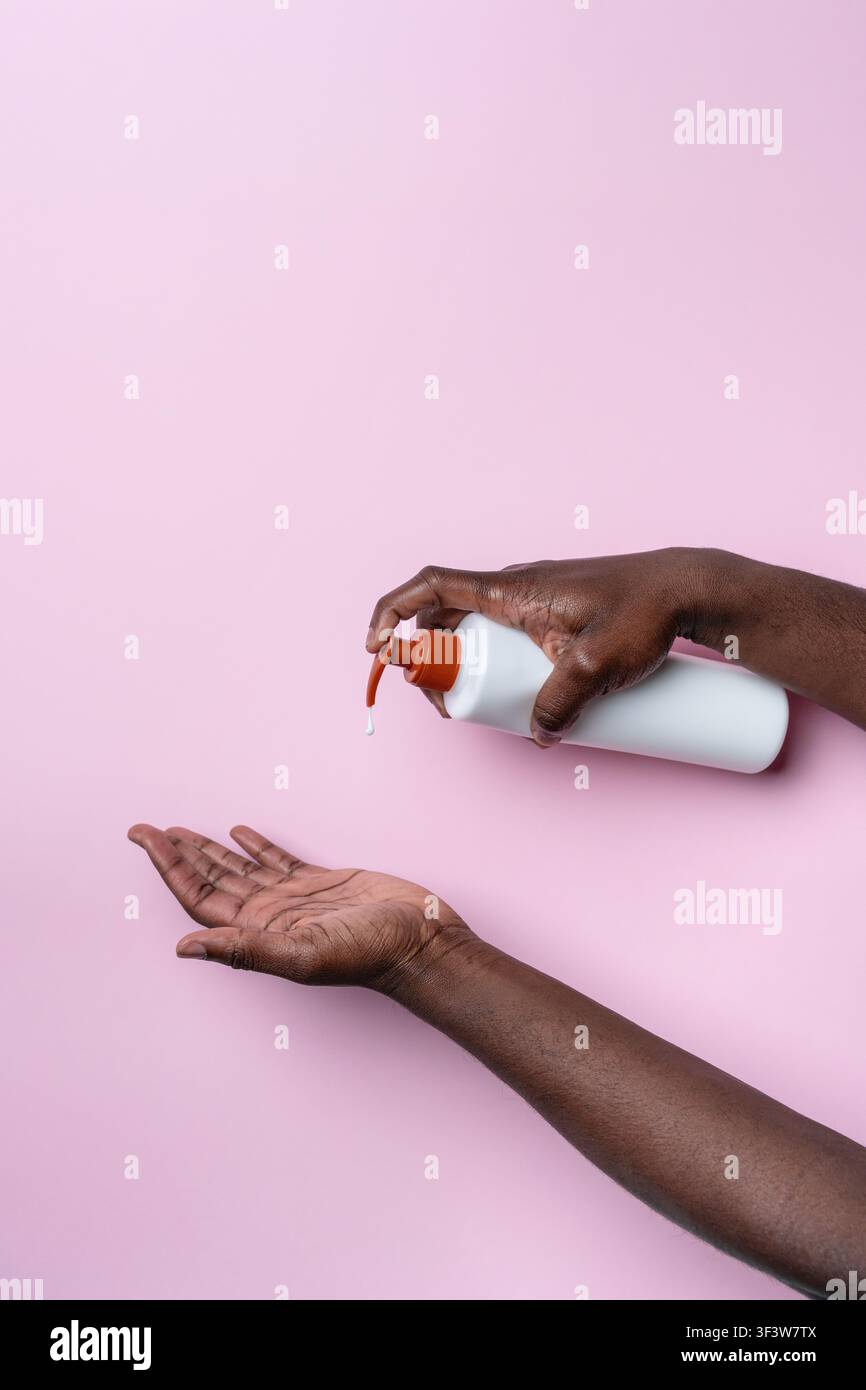 Prima di mani di un uomo nero che eroga sapone liquido da una bottiglia a pompa nella palma aperta su uno sfondo rosa, evidenziando l'igiene, la routine di cura della pelle, un Foto Stock