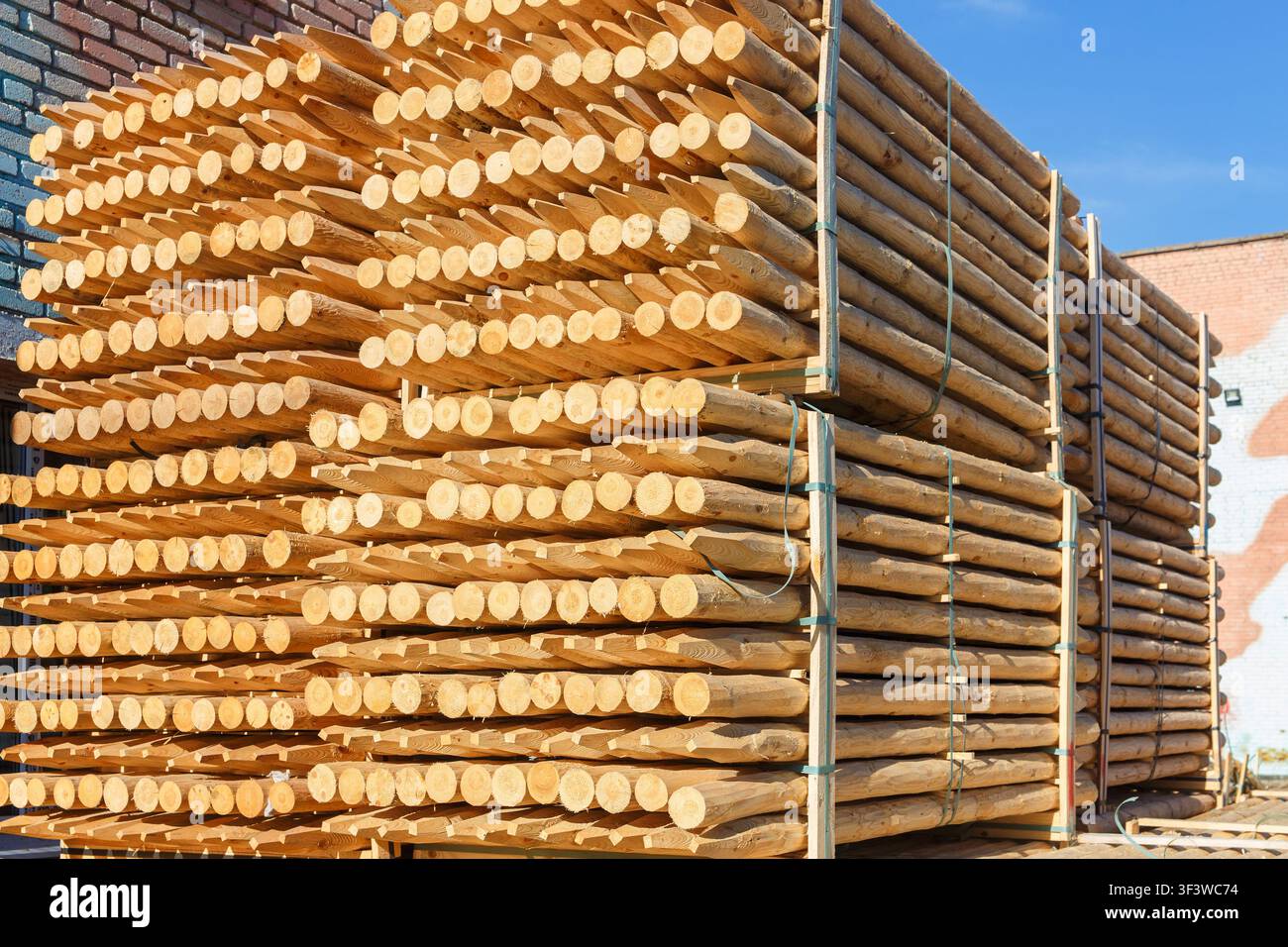 Pile di pali di legno appuntiti legati con cinghie conservati all'aperto vicino a un muro di mattoni sotto un cielo blu, legname per recinzioni e costruzioni. Foto Stock