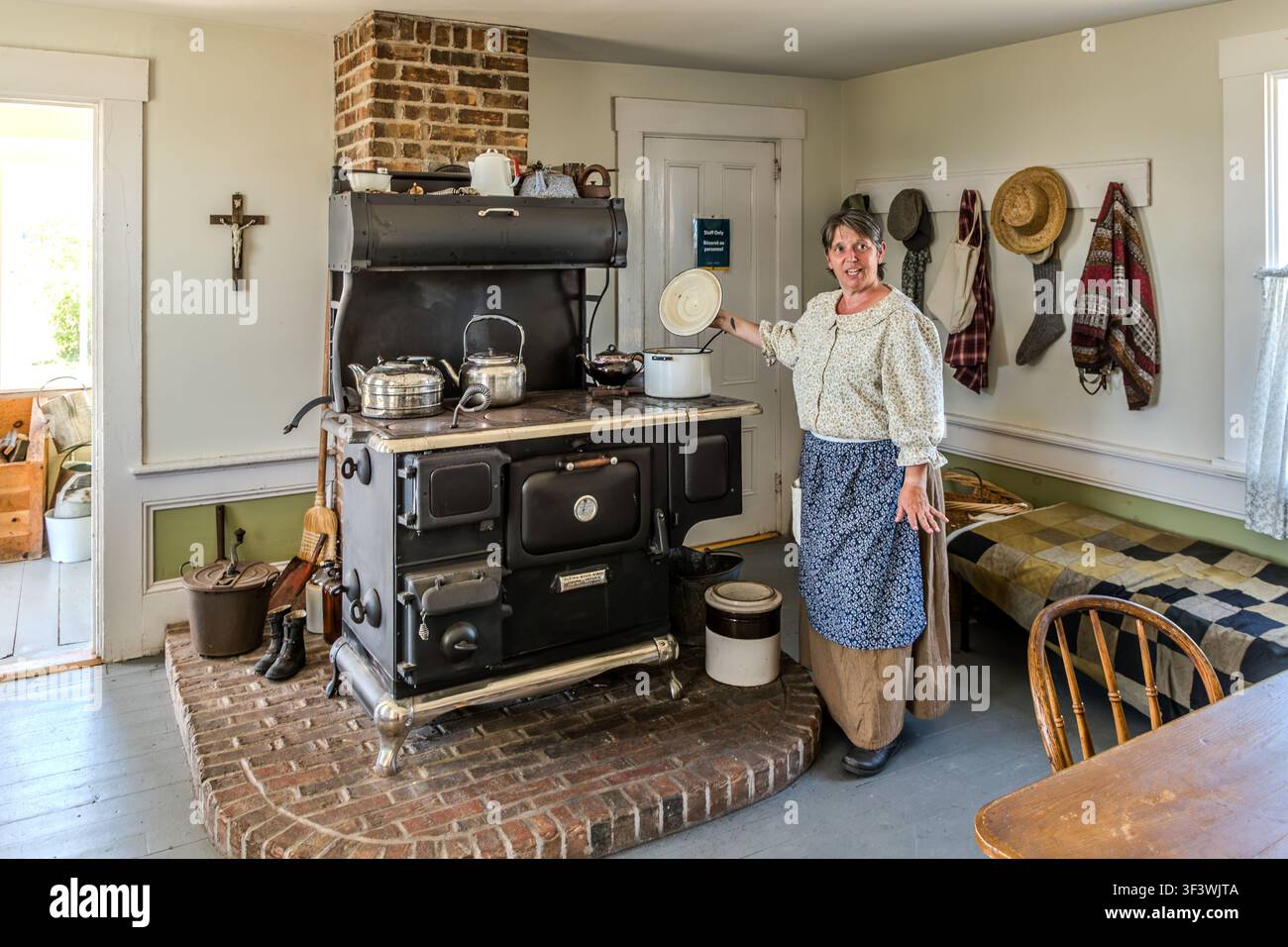 Un'attrice di rievocazione si trova accanto al fornello in ghisa in una fattoria storica al Le Village historique acadien de la Nouvelle-Écosse su Old Church Road a Lower West Pubnico, nella Nuova Scozia. Spiega ai visitatori com'era la vita quotidiana in una cucina acadiana intorno al 1900, con pentole di rame, brocche di terracotta e vestiti di tutti i giorni appesi al muro. Argyle Municipal, Nuova Scozia, Canada Foto Stock