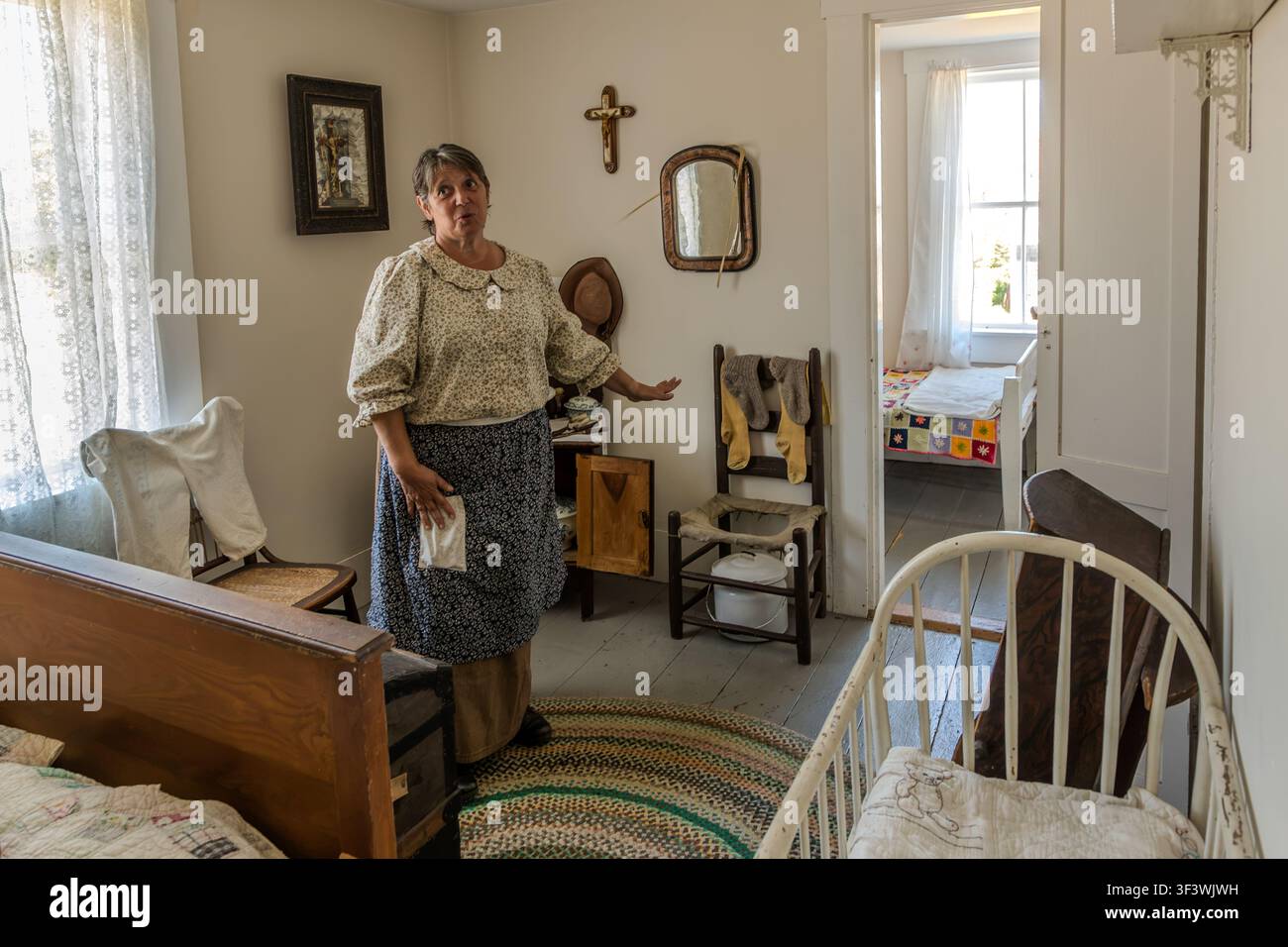 In una camera da letto della fattoria storica del villaggio storico acadiano della Nuova Scozia su Old Church Road a Lower West Pubnico, Nuova Scozia, un’attrice in costume d’epoca spiega la vita quotidiana di una famiglia acadiana intorno al 1900. Gli arredi includono un crocifisso, un tappeto tessuto e una coperta cucita a mano sul letto nella stanza adiacente. Argyle Municipal, Nuova Scozia, Canada Foto Stock