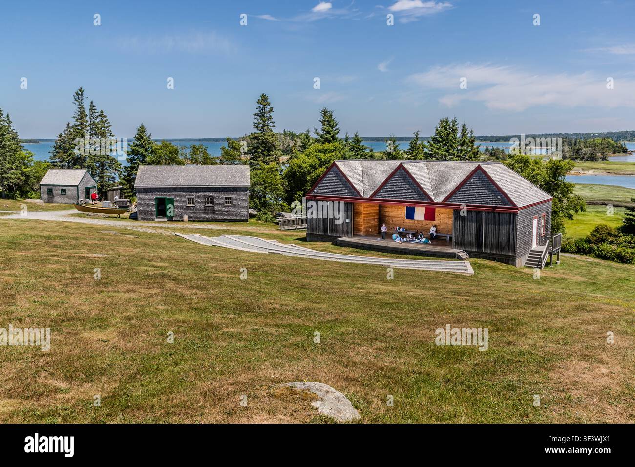 Una vista sui terreni del Villaggio Storico Accadico della Nuova Scozia su Old Church Road a Lower West Pubnico, Nuova Scozia, con un palco all’aperto dove sventola la bandiera acadica e diversi edifici storici sullo sfondo del porto di Pubnico. Alcuni visitatori sono seduti all’ombra dei frontoni del palco. Argyle Municipal, Nuova Scozia, Canada Foto Stock