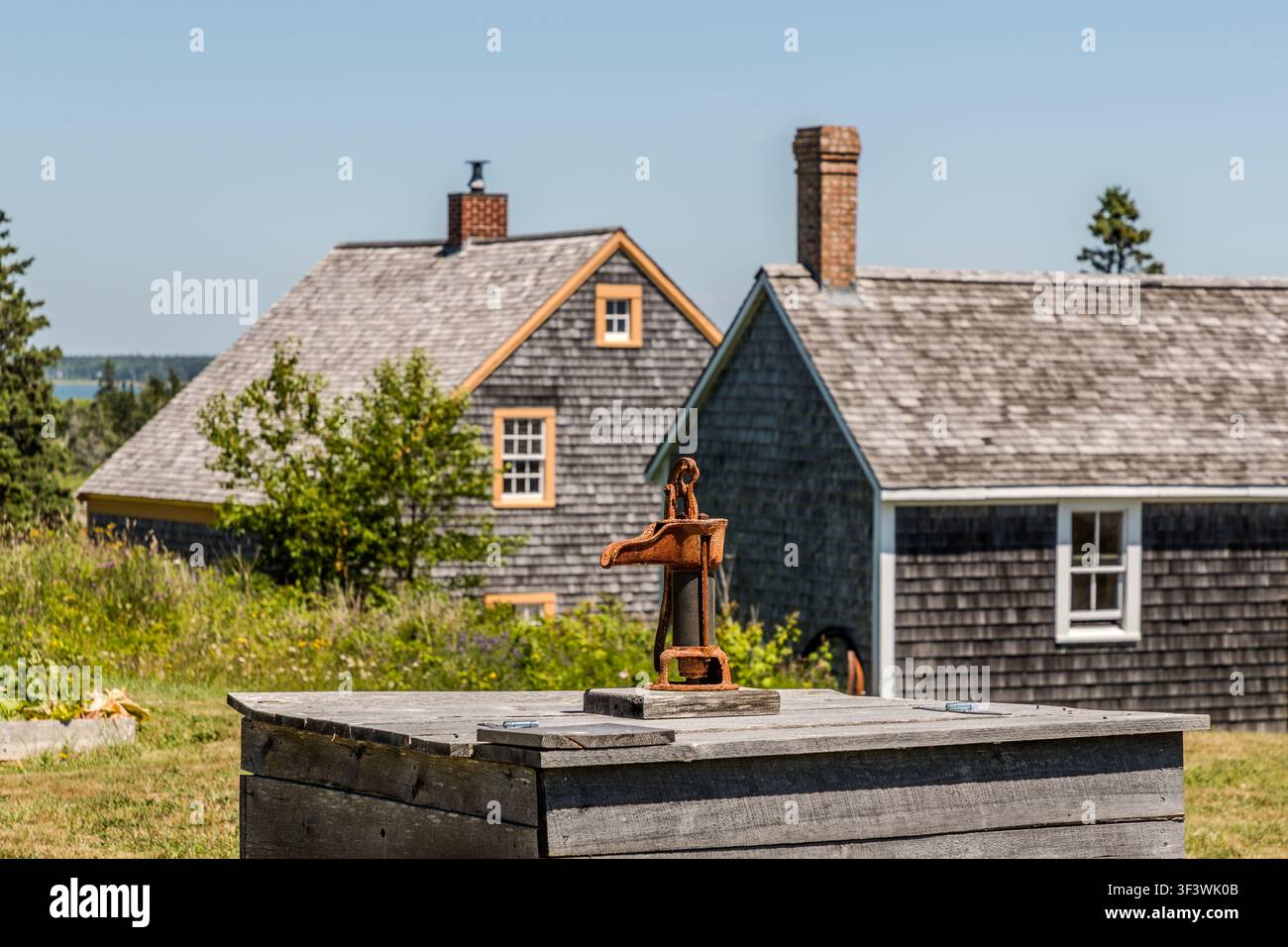 Una pompa manuale arrugginita si trova su una scatola di legno davanti a due case con facciate rivestite di scandole nell'Acadian Historic Village della Nuova Scozia a Lower West Pubnico, Nuova Scozia. Argyle Municipal, Nuova Scozia, Canada Foto Stock