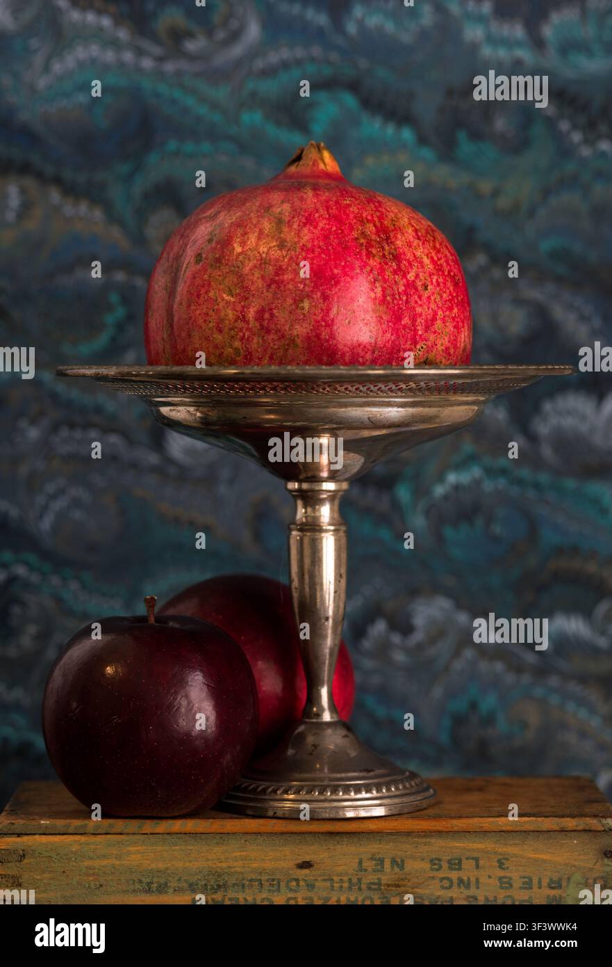 Set di melagrane su una tazza d'argento, con mele rosso scuro su scatole di legno consumate da supermercato sullo sfondo di carta marmorizzata veneziana blu e bianca Foto Stock