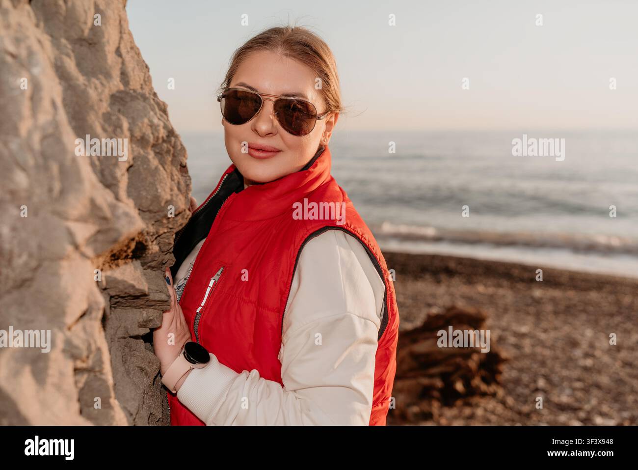 Donna con occhiali da sole da spiaggia che indossa un giubbotto rosso e posa vicino a una grande roccia su una spiaggia di ciottoli durante una tranquilla vacanza al tramonto Foto Stock