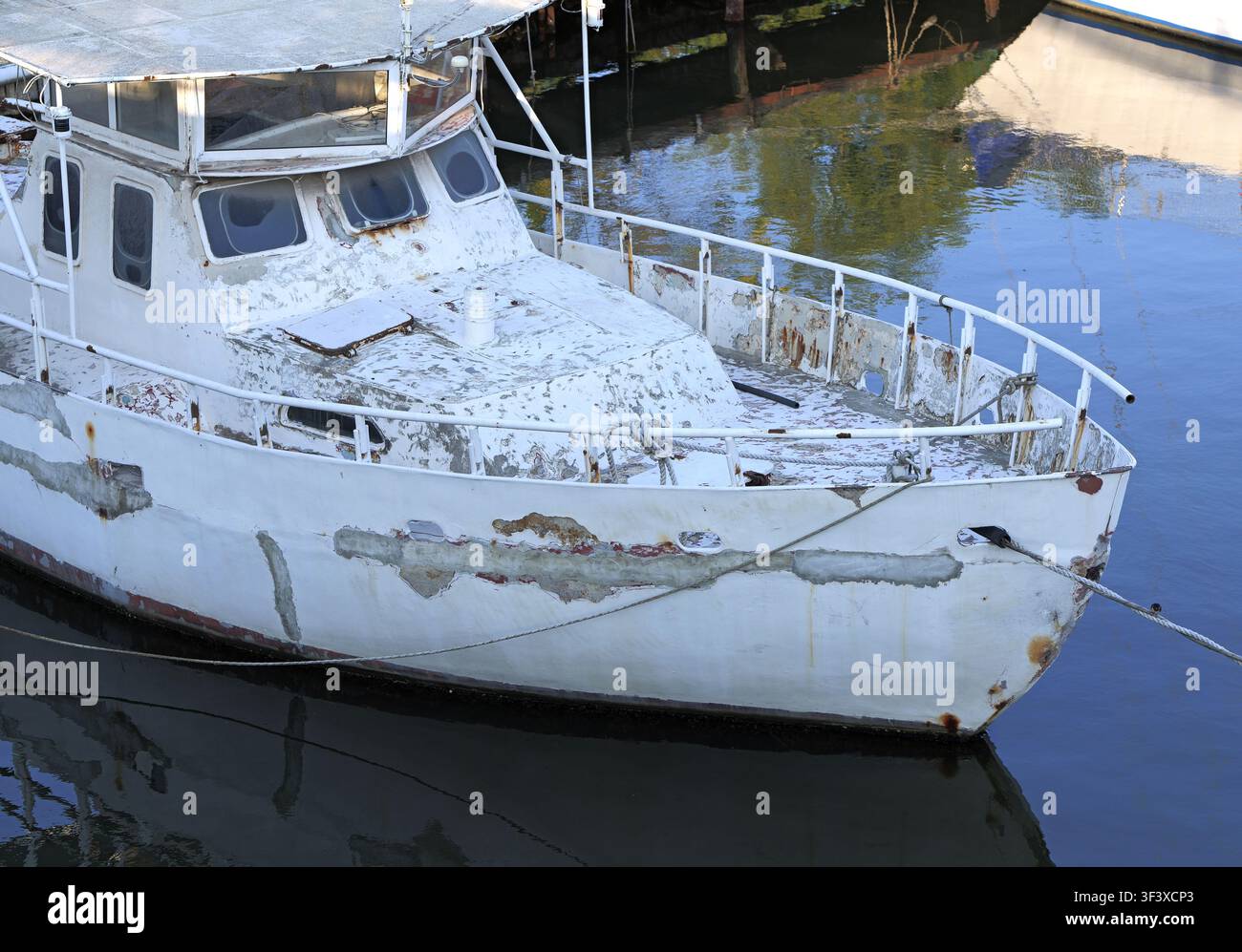 Ex barca da pesca decadente ormeggiata in una tranquilla marina, con vernice bianca scolorita che si stacca dallo scafo, macchie di ruggine e superfici consumate che riflettono Foto Stock