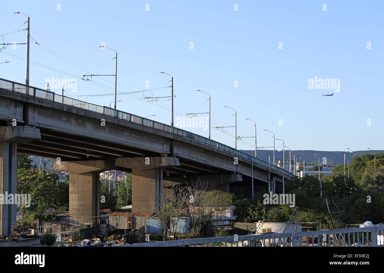 Ponte in cemento con lampioni e cavi che passano sopra un'area urbana dove sono visibili alberi e rifugi improvvisati, con un aereo che vola nel Foto Stock