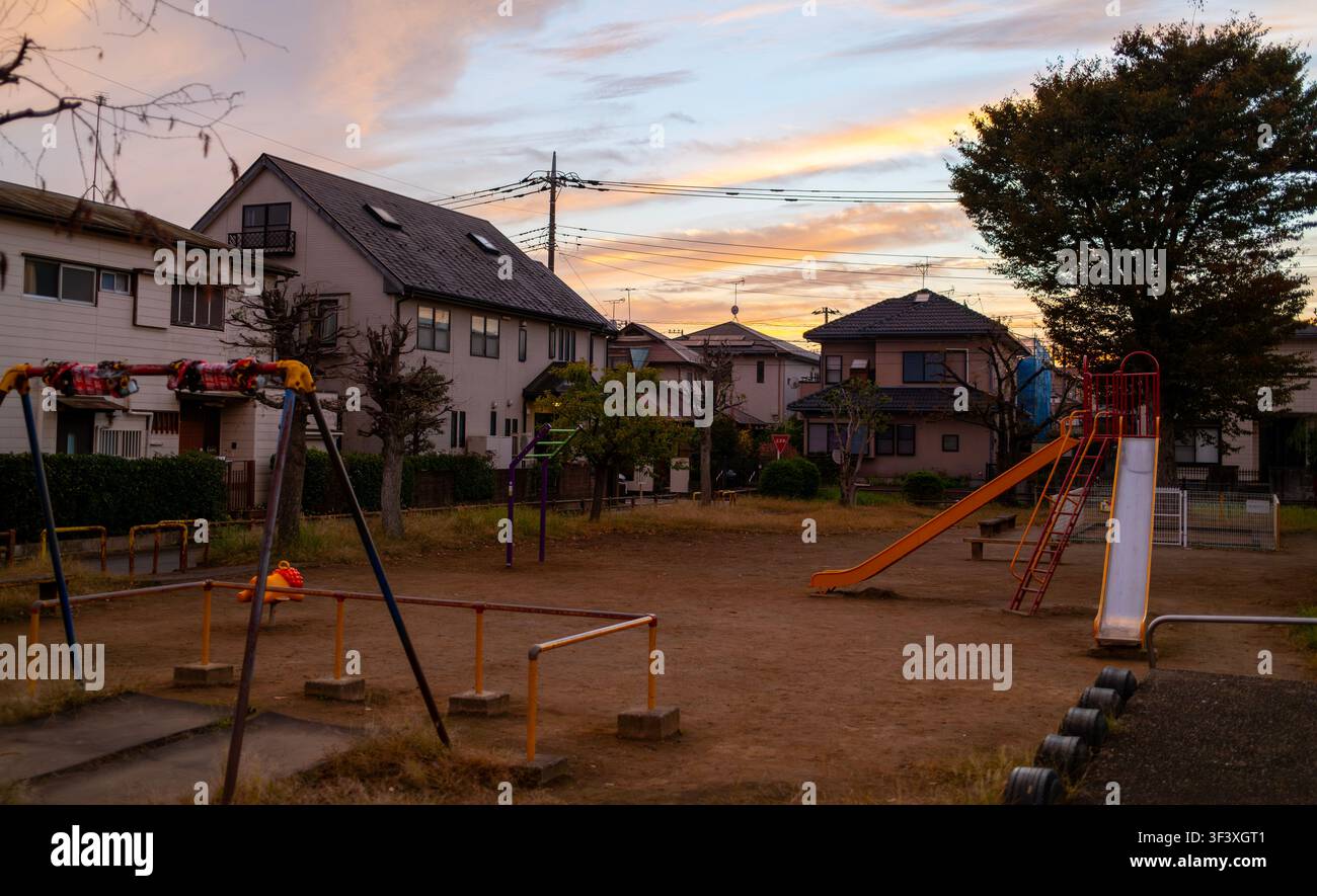 Kasukabe, Giappone – 29 ottobre 2025: Un tranquillo parco giochi di periferia con altalene e scivoli nostalgici, sullo sfondo di un caldo tramonto e case locali. Foto Stock