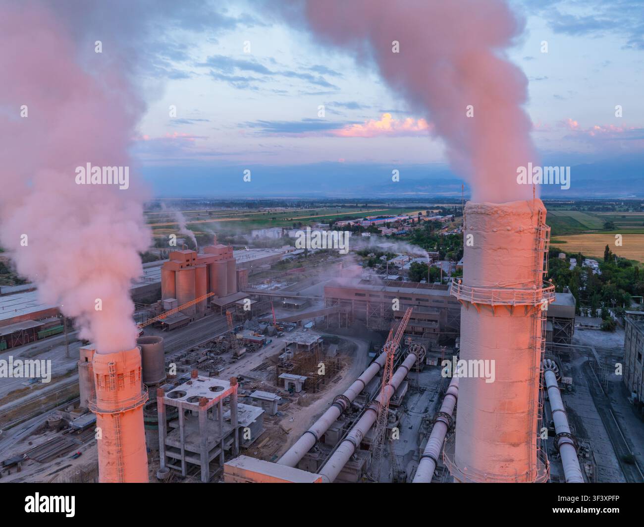 Veduta aerea che cattura un complesso industriale con camini e pennacchi di fumo. Una luce soffusa crea un'atmosfera nebbiosa sulle strutture di produzione e sull'agricoltura Foto Stock
