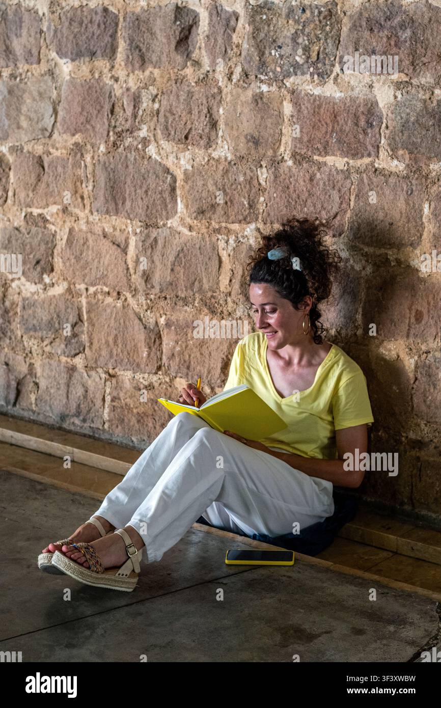 Donna che scrive in un taccuino giallo sul pavimento accanto a un muro di pietra Foto Stock