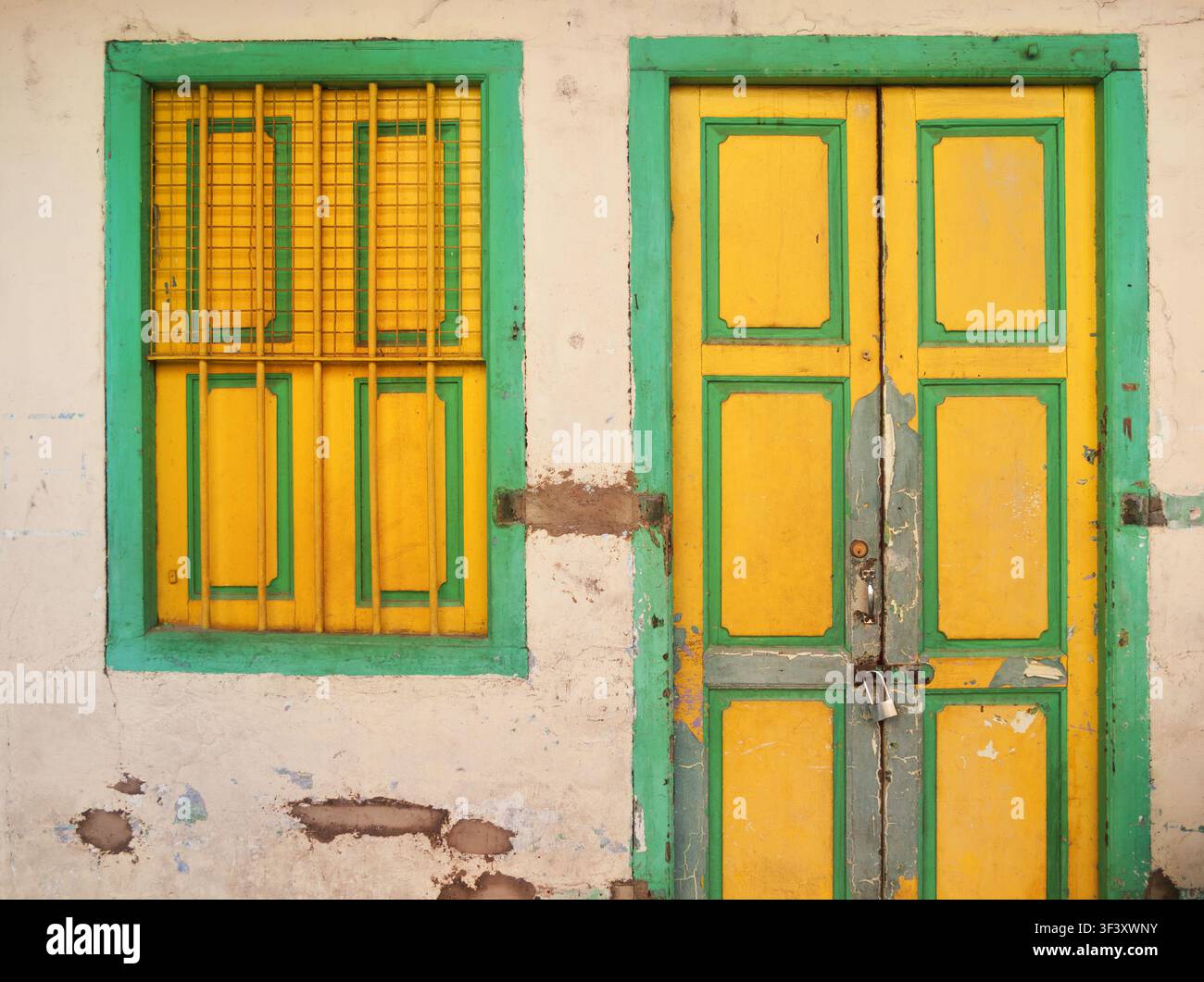 Porta chiusa a chiave gialla e verde abbinata con finestra sbarrata che risalta contro un muro di intonaco consumato e scheggiato Foto Stock