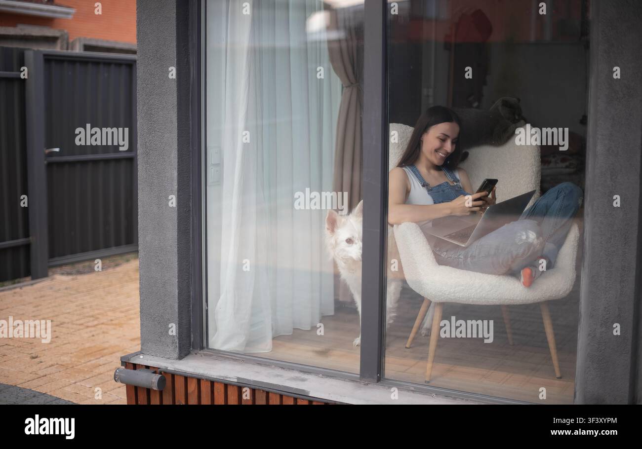 Vista dall'esterno attraverso una grande finestra di una giovane donna sorridente che usa uno smartphone accanto a un laptop, cane bianco e gatto grigio all'interno di una casa moderna. Foto Stock