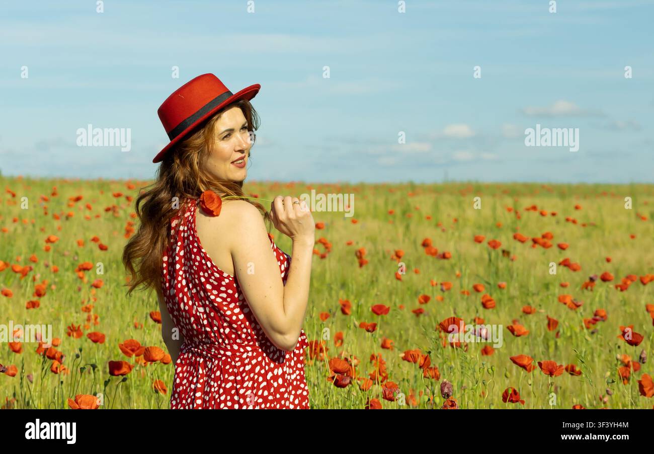 Bella donna dai capelli rossi che guarda indietro con un sorriso gentile mentre indossa un cappello rosso e un vestito rosso in un campo vibrante di papaveri e orzo sotto un cielo blu. Foto Stock