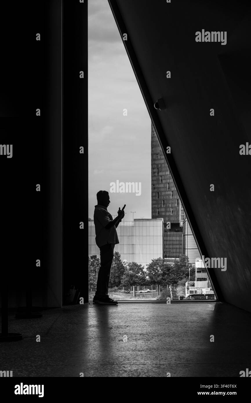 Un uomo d'affari in silhouette utilizza uno smartphone da una cornice di cemento brutalista, catturando lo skyline urbano e l'edificio giallo oltre. Foto Stock
