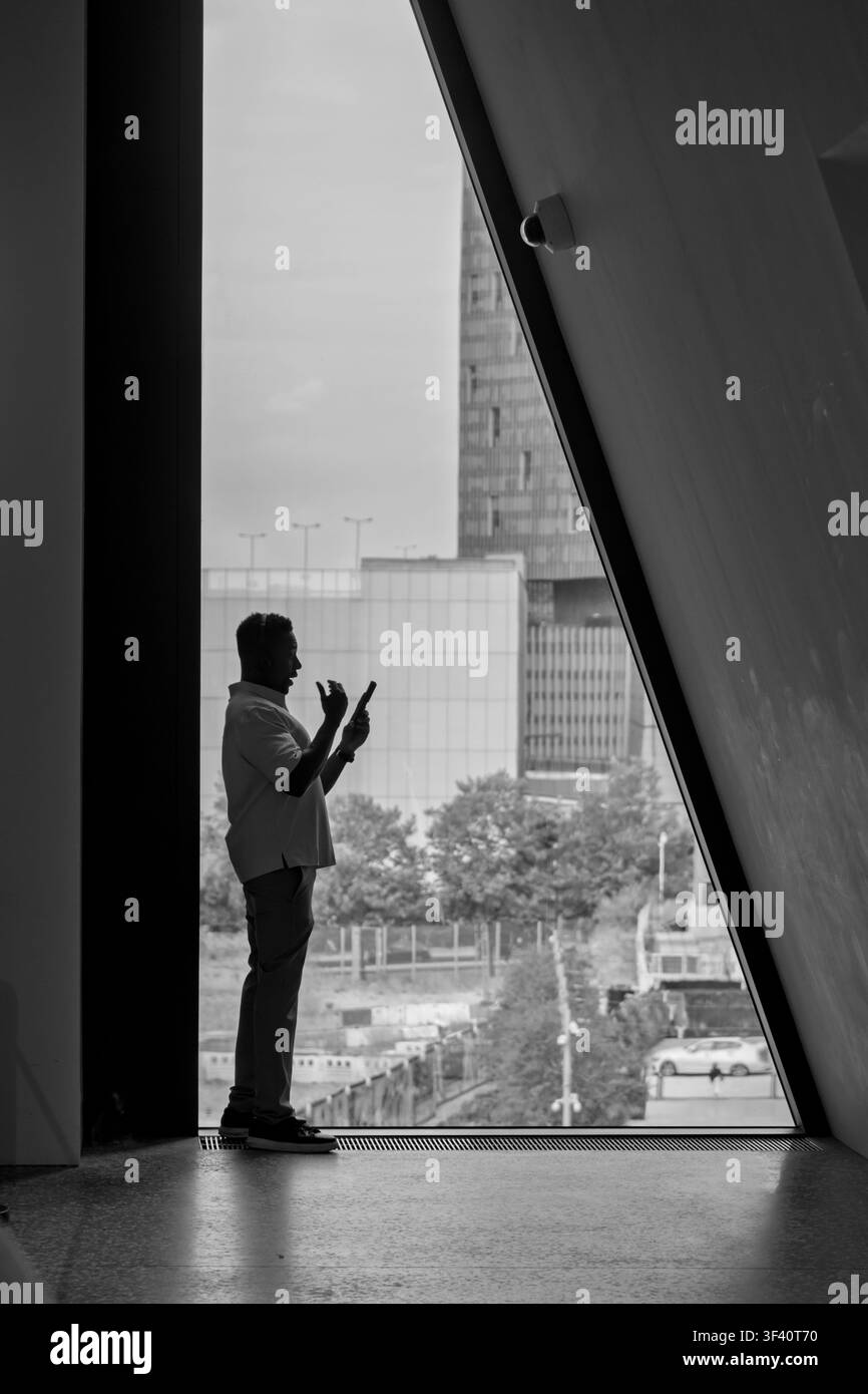 Un uomo d'affari in silhouette utilizza uno smartphone da una cornice di cemento brutalista, catturando lo skyline urbano e l'edificio giallo oltre. Foto Stock
