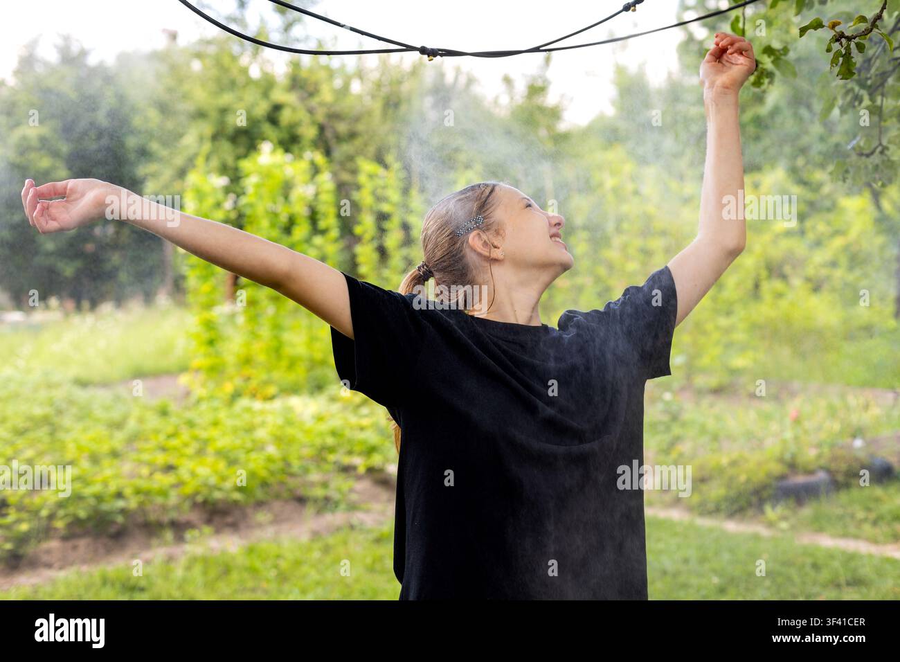 Giovane donna che godette di una nebbia rinfrescante nel giardino all'aperto. Una giovane donna sorride con le braccia alzate mentre gode di una leggera nebbia rinfrescante in un lussureggiante giardino estivo. Foto Stock