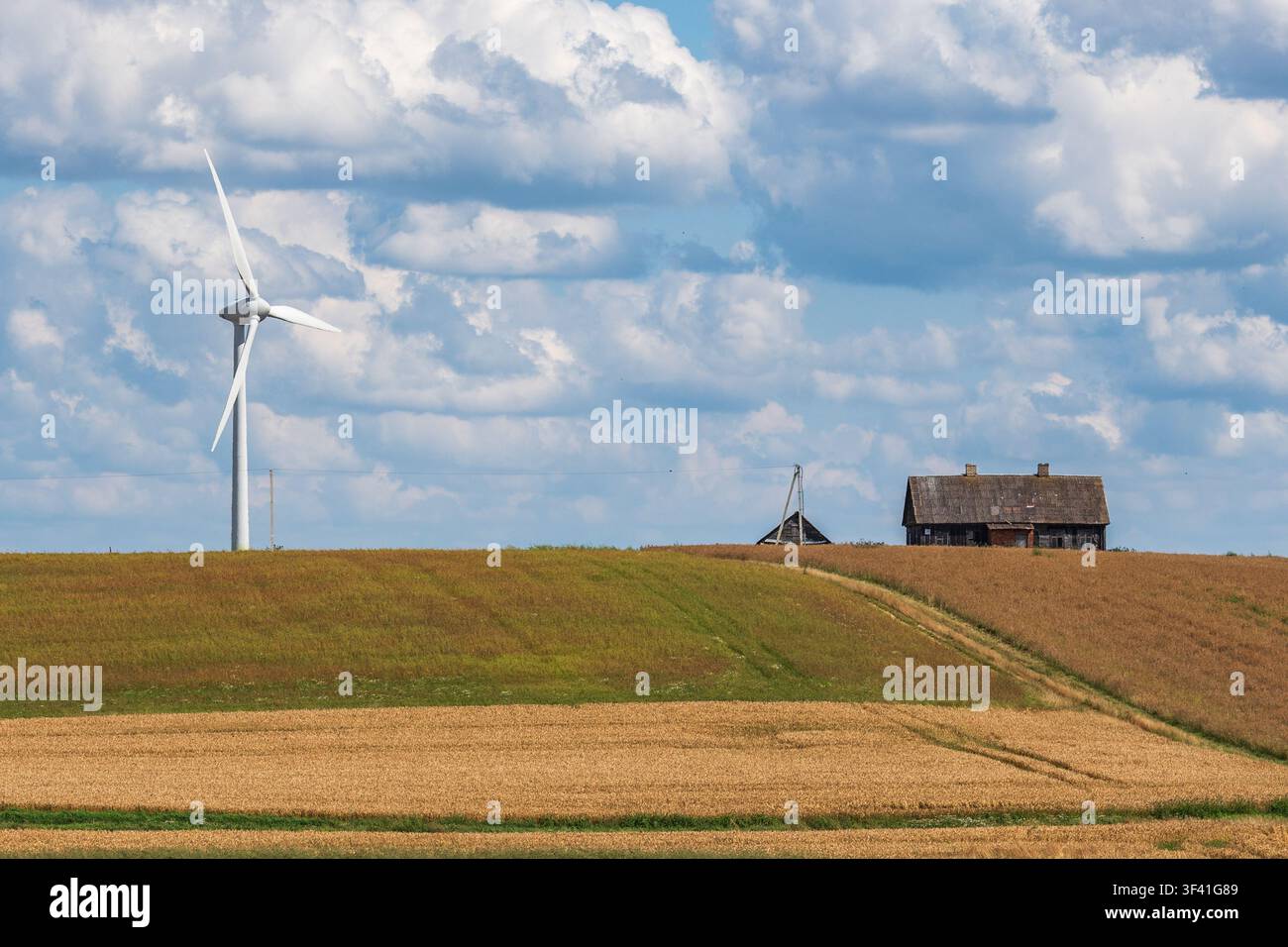 Turbolino eolico moderno posizionato su un terreno agricolo ondulato con una fattoria o un fienile in legno, paesaggio sotto un cielo parzialmente nuvoloso Foto Stock