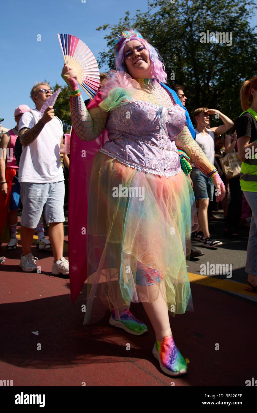 BERLINO, GERMANIA, 25 luglio 2026: Un partecipante sorridente che indossa una gonna di tulle colorata e un corsetto arcobaleno tiene un ventaglio alla parata del CSD di Berlino. Foto Stock