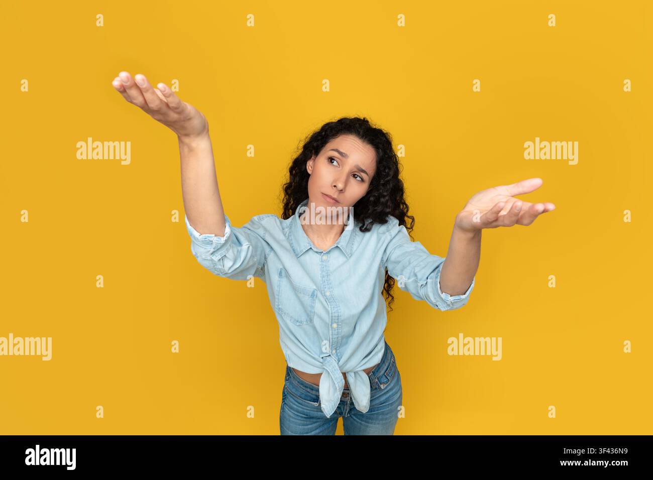 Giovane donna pensierosa con capelli ricci che indica uno sfondo giallo, si sente confusa Foto Stock